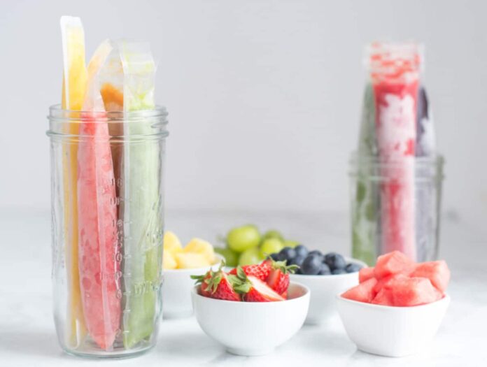 Two glass jars of homemade fruit popsicles sit on a counter alongside small white dishes of fruit.